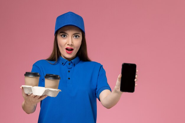 Mensajero femenino de vista frontal en uniforme azul y capa sosteniendo tazas de café de entrega y teléfono en la pared rosa