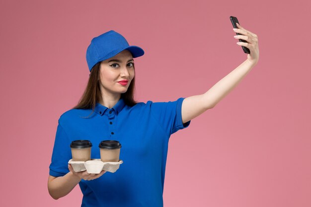 Mensajero femenino de vista frontal en uniforme azul y capa sosteniendo tazas de café de entrega tomando una foto en la pared rosa