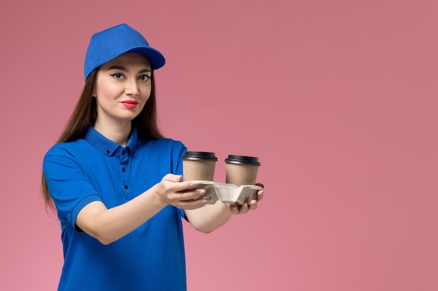 Mensajero femenino de vista frontal en uniforme azul y capa sosteniendo tazas de café de entrega en trabajador de pared rosa