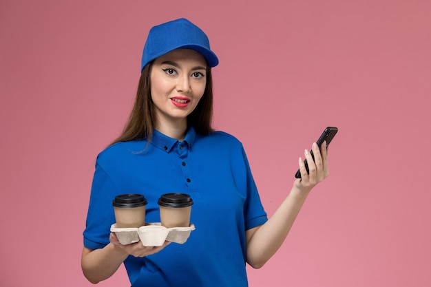 Mensajero femenino de vista frontal en uniforme azul y capa sosteniendo tazas de café de entrega usando su teléfono en la pared rosa