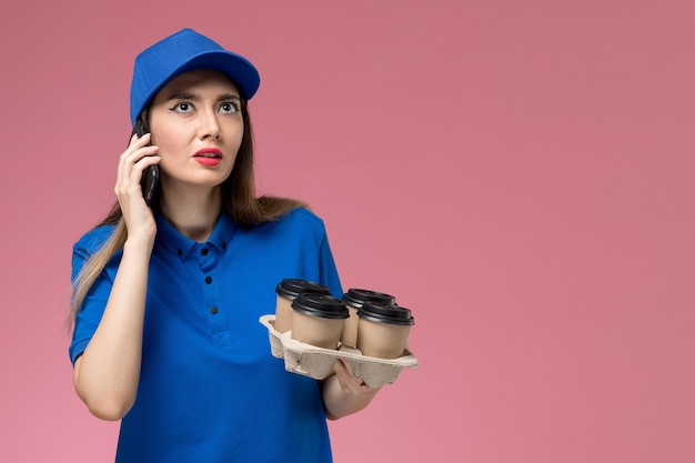 Mensajero femenino de vista frontal en uniforme azul y capa sosteniendo tazas de café hablando por teléfono en la pared rosa