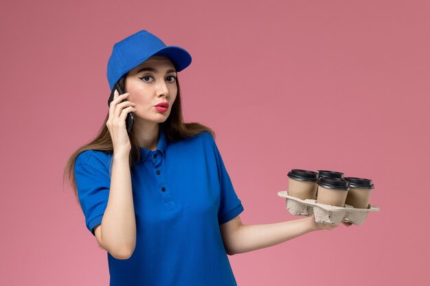 Mensajero femenino de vista frontal en uniforme azul y capa sosteniendo tazas de café hablando por teléfono en la pared rosa