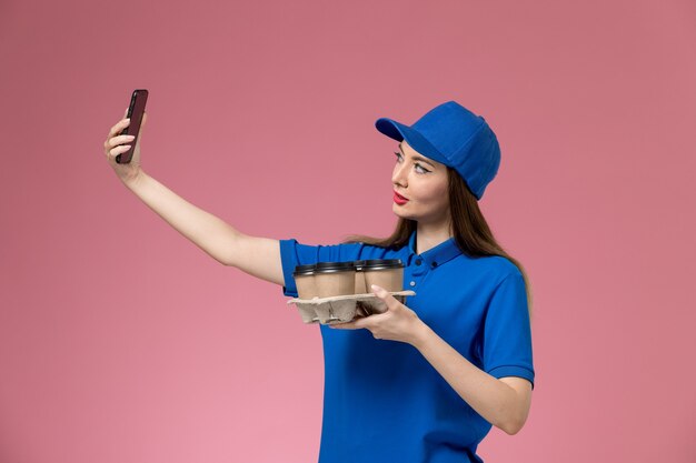 Mensajero femenino de vista frontal en uniforme azul y capa sosteniendo tazas de café tomando una foto en el trabajo de pared rosa