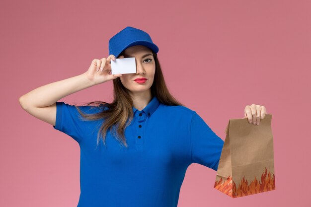 Mensajero femenino de vista frontal en uniforme azul y capa con tarjeta blanca y paquete de comida de papel en la pared rosa