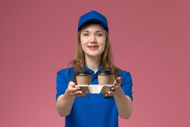 Mensajero femenino de vista frontal en uniforme azul entregando tazas de café marrón en uniforme de servicio de escritorio rosa entrega de trabajo de la empresa