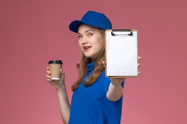 Mensajero femenino de la vista frontal en uniforme azul que sostiene la taza de café marrón con el bloc de notas y una sonrisa en el uniforme de trabajo de servicio de fondo rosa claro que entrega la empresa