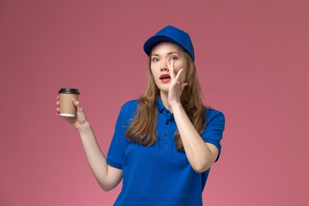 Mensajero femenino de la vista frontal en uniforme azul que sostiene la taza de café marrón en la empresa de entrega de trabajo uniforme de trabajo de servicio de escritorio rosa claro