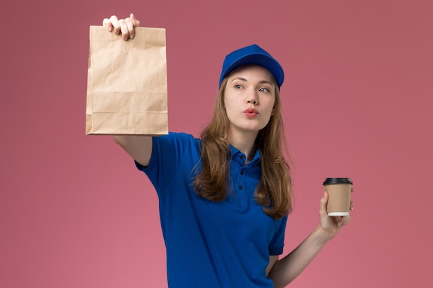 Mensajero femenino de la vista frontal en uniforme azul que sostiene la taza de café marrón con el paquete de alimentos en la empresa de entrega de uniforme de servicio de fondo rosa claro