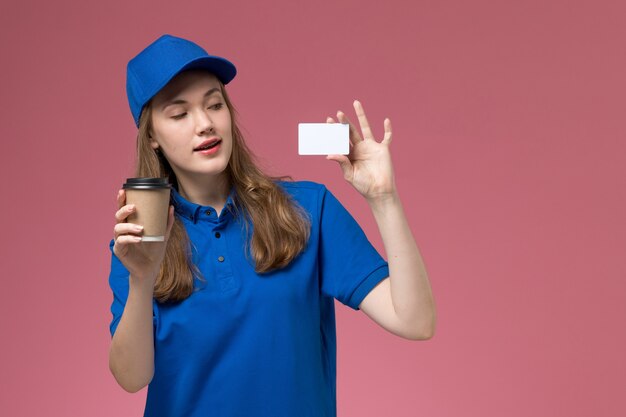 Mensajero femenino de la vista frontal en uniforme azul que sostiene la taza de café marrón con la tarjeta blanca en la empresa de entrega de trabajo uniforme de trabajo de servicio de fondo rosa