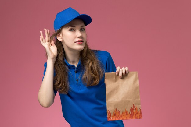 Mensajero femenino de vista frontal en uniforme azul sosteniendo el paquete de alimentos tratando de escuchar en la empresa de uniforme de servicio de trabajador de trabajo de escritorio rosa