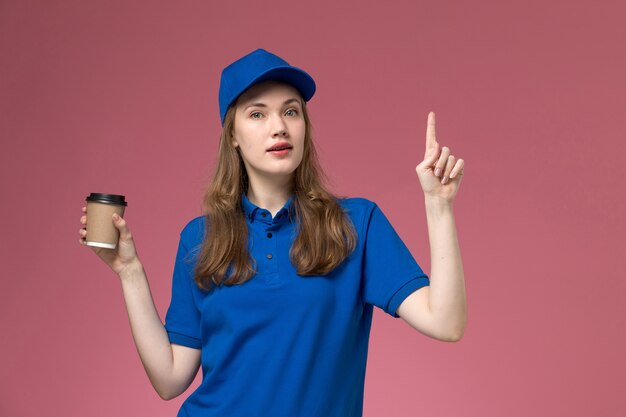Mensajero femenino de vista frontal en uniforme azul sosteniendo una taza de café marrón con el dedo levantado en la empresa de entrega de uniforme de trabajo de servicio de escritorio rosa claro
