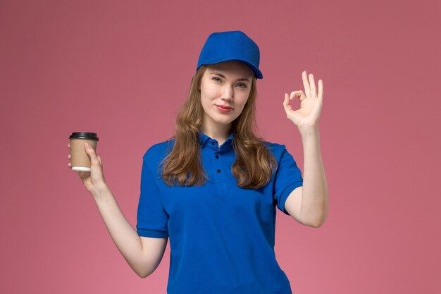 Mensajero femenino de vista frontal en uniforme azul sosteniendo una taza de café marrón que muestra bien el cartel en el uniforme de servicio de escritorio rosa claro que entrega el trabajo de la empresa