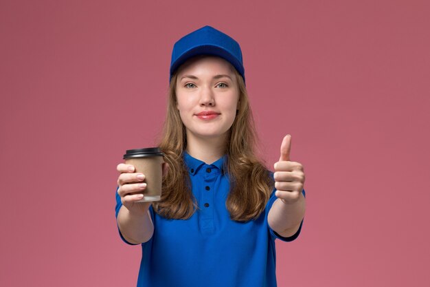 Mensajero femenino de vista frontal en uniforme azul sosteniendo una taza de café marrón sonriendo en uniforme de servicio de escritorio rosa entrega trabajo de trabajo de la empresa