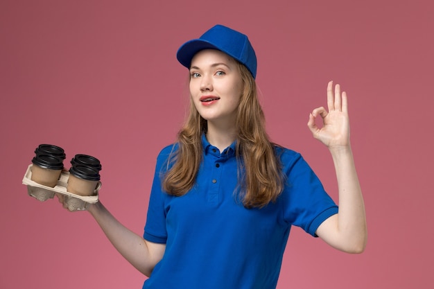 Mensajero femenino de vista frontal en uniforme azul sosteniendo tazas de café de entrega marrón mostrando bien signo en el servicio de escritorio rosa trabajador de la empresa uniforme