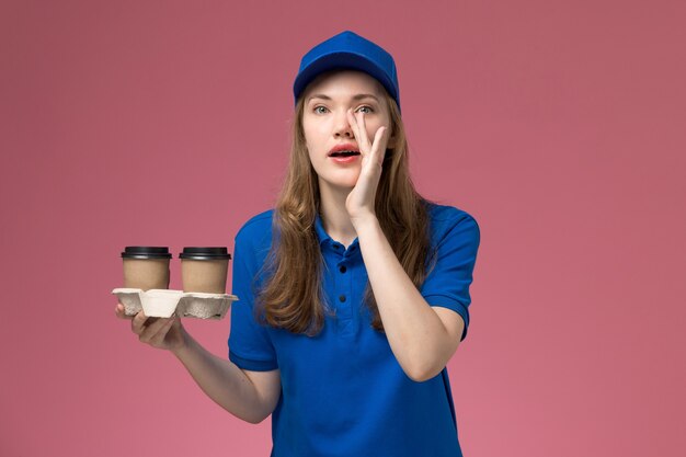 Mensajero femenino de vista frontal en uniforme azul sosteniendo tazas de café de entrega marrón susurrando sobre el uniforme de servicio de escritorio rosa entrega trabajo de la empresa