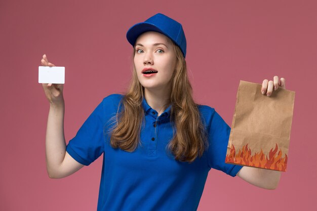 Mensajero femenino de vista frontal en uniforme azul con tarjeta blanca y paquete de comida con expresión de sorpresa en el trabajo de la empresa uniforme de servicio de escritorio de luz rosa