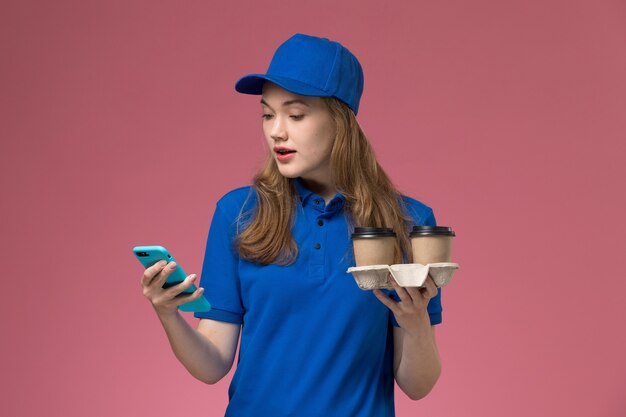 Mensajero femenino de vista frontal en uniforme azul usando su teléfono sosteniendo tazas de café de entrega marrón en empresa de uniforme de trabajo de servicio de escritorio rosa