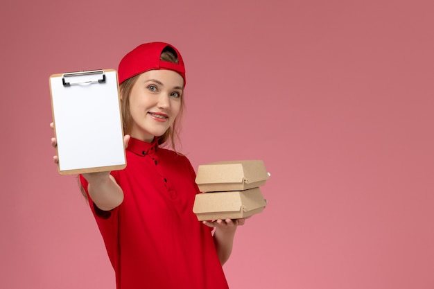 Mensajero femenino de vista frontal en uniforme rojo y capa con bloc de notas y pequeños paquetes de comida de entrega sobre fondo rosa claro trabajador de entrega uniforme