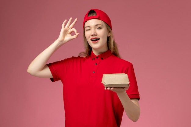 Mensajero femenino de vista frontal en uniforme rojo y capa guiñando un ojo sosteniendo un pequeño paquete de comida de entrega en la pared rosa, trabajo uniforme de la empresa de servicio de entrega