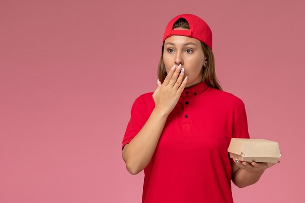 Mensajero femenino de vista frontal en uniforme rojo y capa con paquete de comida de entrega en el fondo rosa empresa de servicio de entrega uniforme trabajo trabajo niña