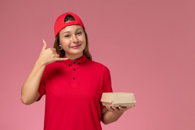 Mensajero femenino de vista frontal en uniforme rojo y capa con paquete de comida de entrega en la pared rosa, chica de trabajo de empresa de servicio de entrega uniforme