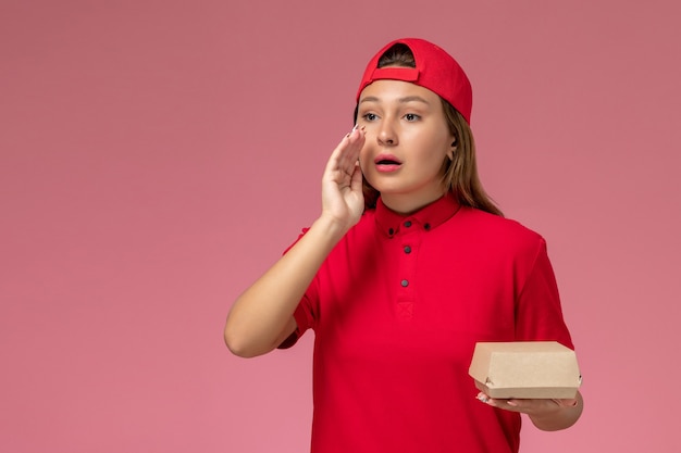 Mensajero femenino de vista frontal en uniforme rojo y capa con paquete de comida de entrega en la pared rosa, trabajador de empresa de servicio de entrega uniforme trabajo niña