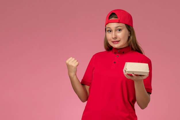 Mensajero femenino de vista frontal en uniforme rojo y capa con paquete de comida de entrega en la pared rosa, trabajador de trabajo de empresa de servicio de entrega uniforme