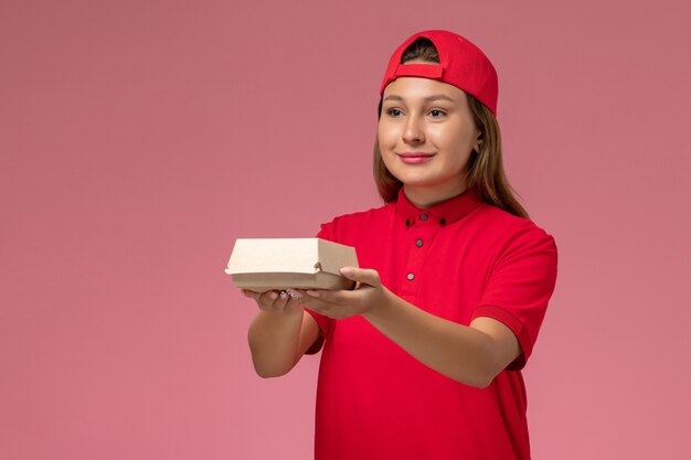 Mensajero femenino de vista frontal en uniforme rojo y capa con paquete de comida de entrega en la pared rosa, trabajo de trabajador de trabajo de empresa de servicio de entrega uniforme