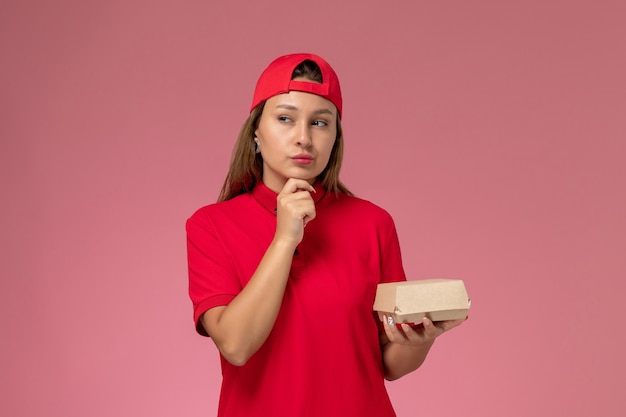 Mensajero femenino de vista frontal en uniforme rojo y capa con paquete de comida de entrega pensando en la pared rosa, trabajo de empresa de servicio de entrega uniforme