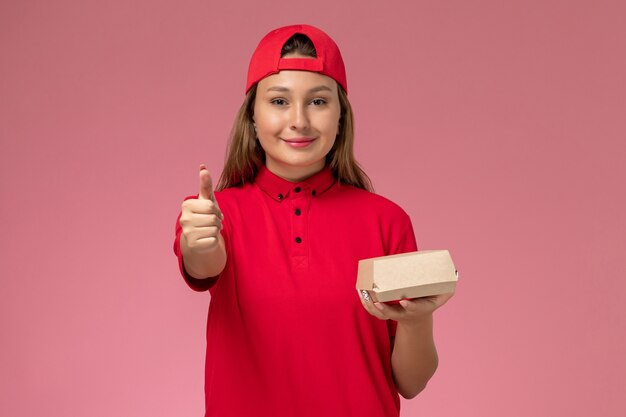 Mensajero femenino de vista frontal en uniforme rojo y capa con paquete de comida de entrega en el trabajo de trabajo de empresa de servicio de entrega uniforme de fondo rosa