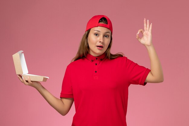 Mensajero femenino de vista frontal en uniforme rojo y capa con paquete de comida de entrega vacío en la pared de color rosa claro, chica de empresa de servicio de entrega uniforme