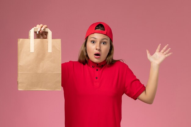 Mensajero femenino de vista frontal en uniforme rojo y capa con paquete de papel de entrega en la pared rosa, trabajo de servicio de entrega uniforme