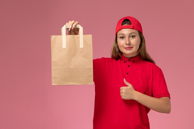 Mensajero femenino de vista frontal en uniforme rojo y capa con paquete de papel de entrega en la pared rosa, trabajo de trabajador de servicio de entrega uniforme de trabajo