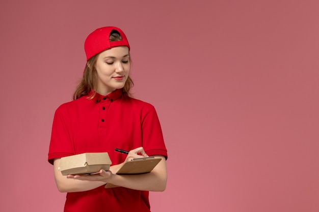 Mensajero femenino de vista frontal en uniforme rojo y capa con pequeño paquete de comida de entrega con bloc de notas escrito en la pared rosa, chica uniforme de servicio de entrega