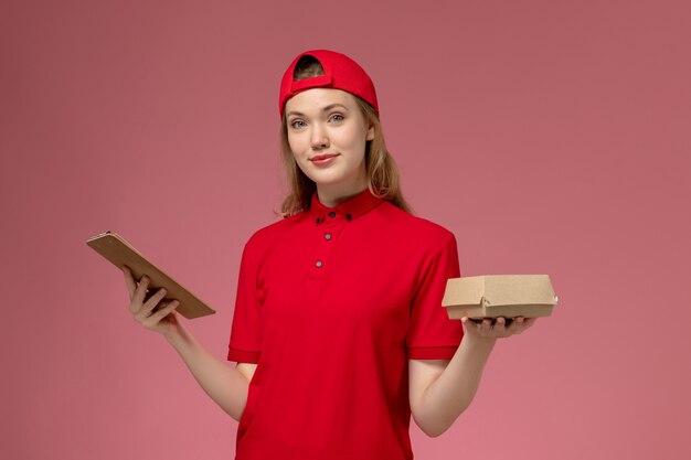 Mensajero femenino de vista frontal en uniforme rojo y capa con pequeño paquete de comida de entrega con bloc de notas en la pared rosa claro, uniforme de trabajo de la empresa de servicios de entrega