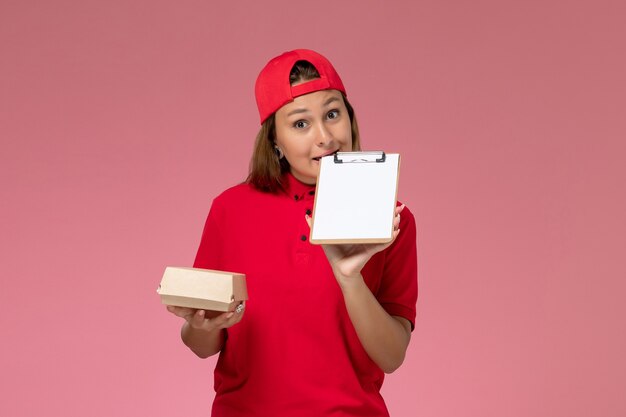 Mensajero femenino de vista frontal en uniforme rojo y capa con pequeño paquete de comida de entrega y bloc de notas en la pared rosa, empresa de servicio de entrega uniforme