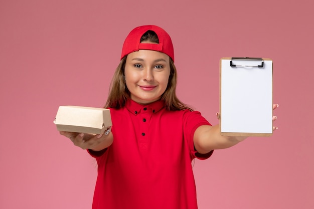 Mensajero femenino de vista frontal en uniforme rojo y capa con pequeño paquete de comida de entrega y bloc de notas en la pared rosa, empresa de servicio de entrega uniforme