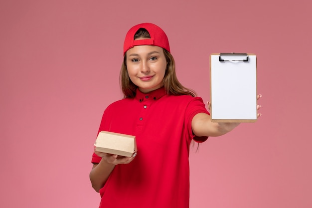 Mensajero femenino de vista frontal en uniforme rojo y capa con pequeño paquete de comida de entrega y bloc de notas en la pared rosa, trabajador de la empresa de servicio de entrega uniforme