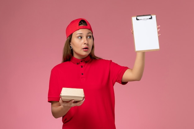 Mensajero femenino de vista frontal en uniforme rojo y capa con pequeño paquete de comida de entrega y bloc de notas en la pared rosa, trabajador de la empresa de servicio de entrega uniforme