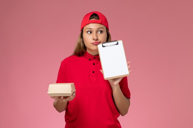 Mensajero femenino de vista frontal en uniforme rojo y capa con pequeño paquete de comida de entrega y bloc de notas pensando en la pared rosa, servicio de entrega uniforme