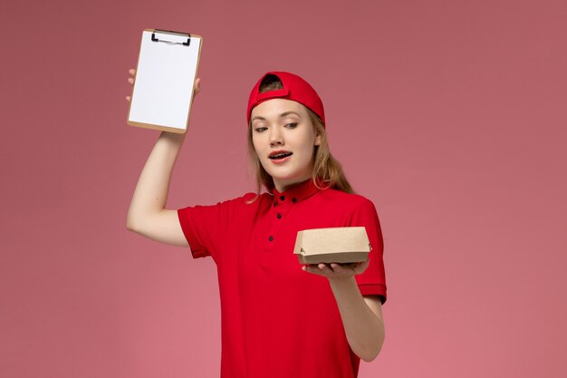 Mensajero femenino de vista frontal en uniforme rojo y capa con pequeño paquete de comida de entrega con bloc de notas en el uniforme de trabajo de la empresa de servicio de entrega de escritorio rosa