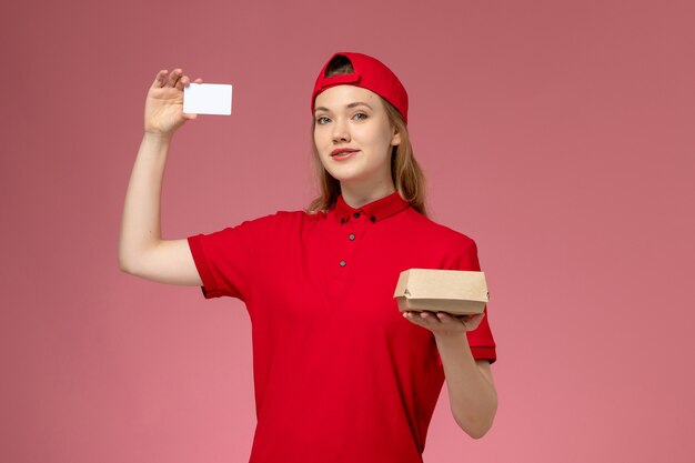 Mensajero femenino de vista frontal en uniforme rojo y capa con pequeño paquete de comida de entrega con tarjeta de plástico blanca en la pared rosa, trabajo de entrega uniforme de servicio