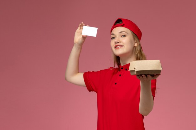 Mensajero femenino de vista frontal en uniforme rojo y capa con pequeño paquete de comida de entrega con tarjeta de plástico blanca en la pared rosa, trabajo de entrega uniforme de servicio