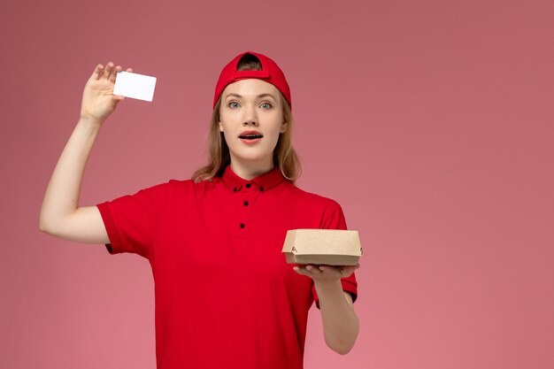 Mensajero femenino de vista frontal en uniforme rojo y capa con pequeño paquete de comida de entrega con tarjeta de plástico blanca en la pared rosa, trabajo de entrega uniforme de servicio