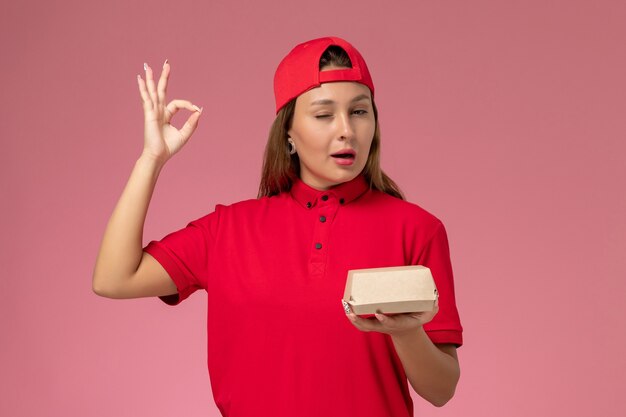 Mensajero femenino de vista frontal en uniforme rojo y capa que sostiene el paquete de comida de entrega en el fondo rosa trabajador de la muchacha del trabajo de la empresa de servicio de entrega uniforme