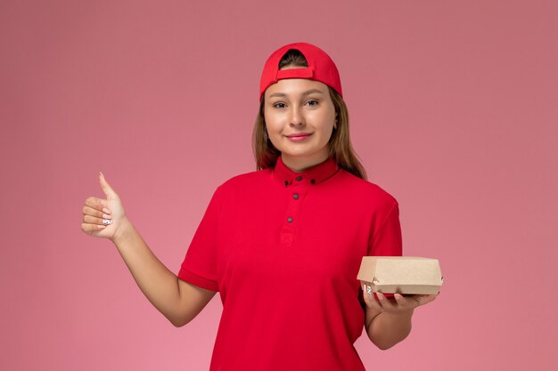Mensajero femenino de la vista frontal en uniforme rojo y capa que sostiene el paquete de comida de entrega en el fondo rosa uniforme de la empresa de entrega de trabajo trabajador chica