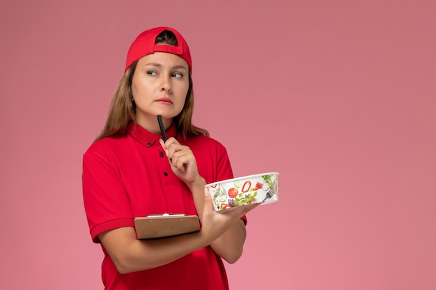 Mensajero femenino de vista frontal en uniforme rojo y capa sosteniendo bloc de notas y cuenco de entrega pensando en el trabajador de trabajo de servicio de entrega uniforme de fondo rosa