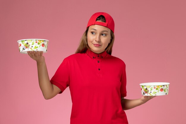 Mensajero femenino de vista frontal en uniforme rojo y capa sosteniendo cuencos de entrega en la pared rosa, servicio de entrega uniforme de trabajador de trabajo