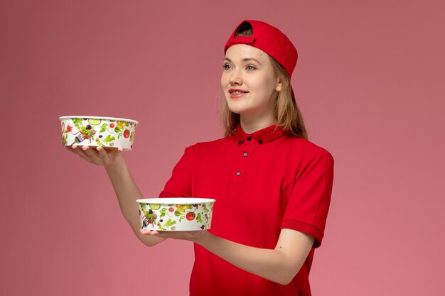 Mensajero femenino de vista frontal en uniforme rojo y capa sosteniendo cuencos de entrega con sonrisa en la pared rosa claro, servicio de entrega uniforme