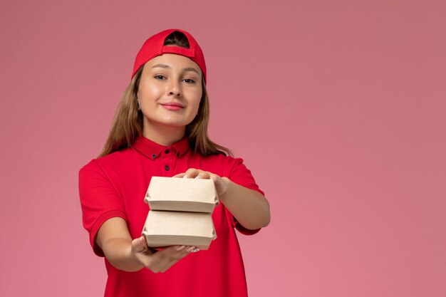 Mensajero femenino de vista frontal en uniforme rojo y capa sosteniendo paquetes de comida de entrega en una pared rosa claro, empresa de servicio de entrega uniforme